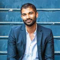 A man with short dark hair and a beard is sitting in front of a blue background. He is wearing a blue blazer over a white shirt, and he is smiling gently at the camera, ready to discuss California Cannabis Law in an upcoming online webinar. CA Norml