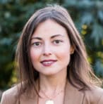 A woman with long, straight brown hair is standing outdoors. She is wearing a brown blazer and a light-colored top, with a pendant necklace. The background features greenery with out-of-focus foliage. She has a neutral expression and looks directly at the camera, as if awaiting her Online Webinar on August 25. CA Norml