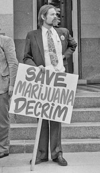 A person in formal attire holds a sign that reads "SAVE MARIJUANA DECRIM" while standing on steps outside a building. The individual has a mustache and long hair. The scene resembles an advocacy moment at the CA State Fair, with part of another person's arm and a doorway visible in the background. CA Norml