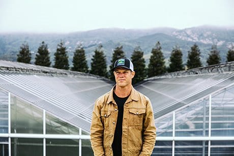 A man wearing a tan jacket and a baseball cap stands in front of greenhouse structures with a backdrop of mountains and fog, reminiscent of the scenic views near the CA State Fair. Pine trees are visible in the distance, adding to the serenity that defines this tranquil setting. CA Norml