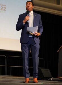 A person in a blue suit stands on a stage holding a microphone and paper. They appear to be speaking to an audience about the program at the CA State Fair, with a screen displaying text and dates in the background. CA Norml