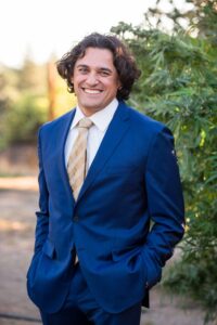 A man stands outdoors smiling, dressed in a blue suit with a beige and white striped tie. He has curly, shoulder-length hair and his hands are in his pockets. The background features green foliage and the vibrant atmosphere of the CA State Fair. CA Norml