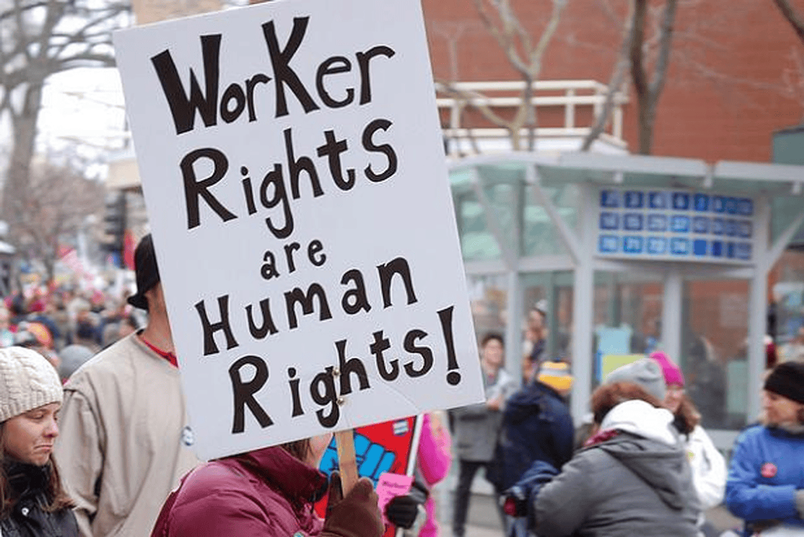 A person is holding a sign that reads "Worker Rights are Human Rights!" at a protest advocating for employment rights. The scene is outdoors with a crowd of people in the background, dressed in winter clothing, and a building with a glass structure is visible. CA Norml