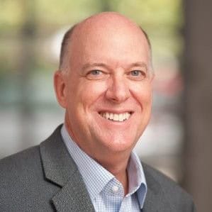 A smiling bald man with light skin, wearing a grey blazer and blue checkered shirt. The blurred background suggests an indoor setting with soft lighting. He is prepared to address the 2024 Legal Seminar hosted by Cal NORML, available for online viewing. CA Norml