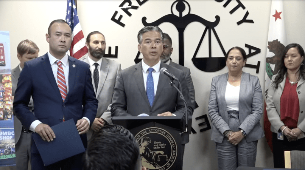 AG Bonta addresses the audience from behind a podium during a press conference, speaking into the microphone about cracking down on illicit cannabis operations through the CAPP Program. Others stand in a semi-circle behind him with serious expressions. An American flag and scales of justice symbol are visible in the background. CA Norml