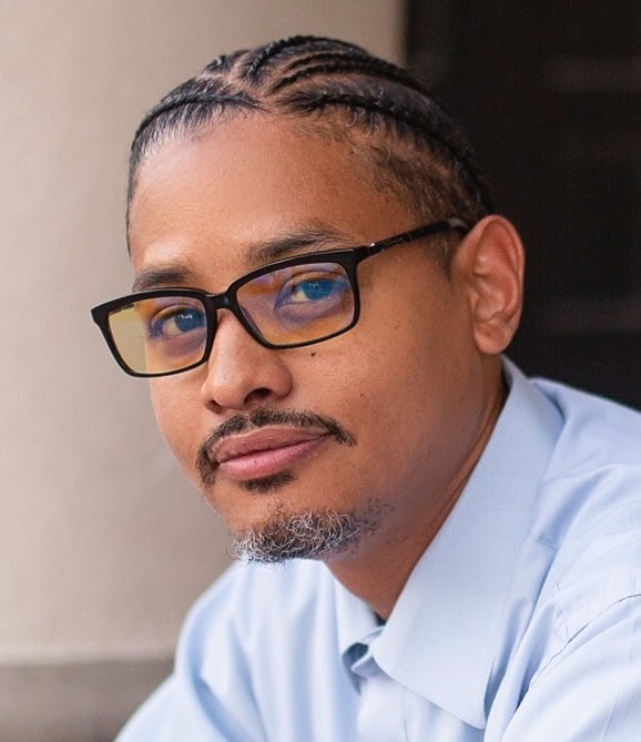 A person with glasses and cornrows gazes confidently at the camera, embodying a professional demeanor. They have a short beard and mustache, and are wearing a light blue dress shirt. The blurred, neutral-toned background suggests a focus on the upcoming Cal NORML 2024 Legal Seminar, which they might attend online. CA Norml