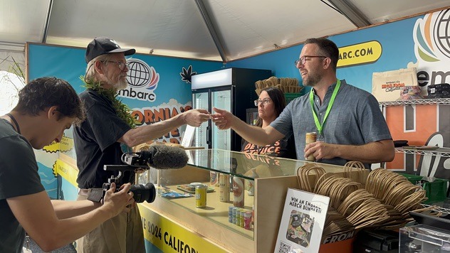 A man behind a booth counter hands a product to a customer at the California State Fair. A woman stands next to him inside the booth, while another man records the interaction with a camera. The booth, highlighting cannabis sales, features colorful banners and shelves with items. CA Norml
