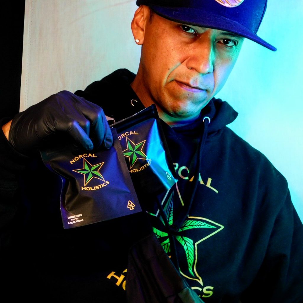 Male wearing Norcal Holistics Hooded Sweatshirt holding cannabis flower bags.