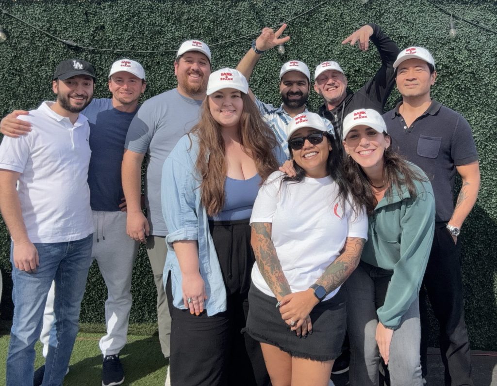 A group of nine people, including the team from Deeproots Partners, smiles and poses outdoors. They are wearing casual clothes, and three individuals sport white caps with red logos. The background features a lush green wall of foliage. CA Norml