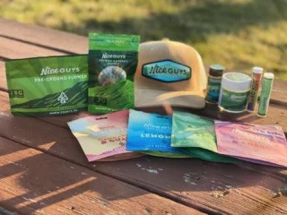 A collection of Nice Guys branded cannabis products from Nice Guys Delivery, including pre-ground flower, edibles, topicals, a hat, and lip balm, displayed on a wooden surface outdoors with grass in the background. CA Norml