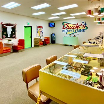 A bright Chuck’s Wellness Center interior features display cases of products, orange seating along the walls, colorful wall art, and a green door. Overhead lights and hanging lamps illuminate the spacious room. CA Norml