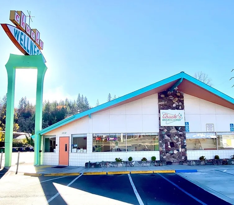 A retro-style diner with a turquoise and pink sign reading "Chuck’s Wellness" stands under a clear blue sky. The building has a slanted roof, large windows, and a stone accent wall, blending nostalgia with the vibe of a modern wellness center. CA Norml