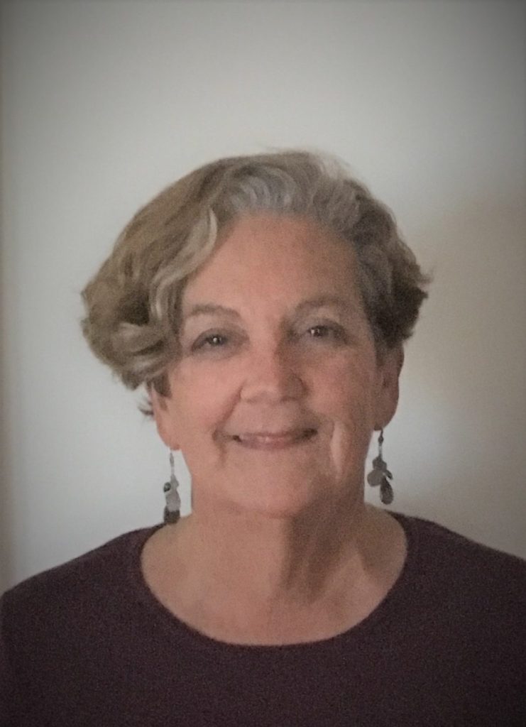 Denise Martellacci, an older woman with short, wavy gray hair, smiles gently at the camera. She is wearing dangling earrings and a dark top, standing against a plain, light-colored background. CA Norml