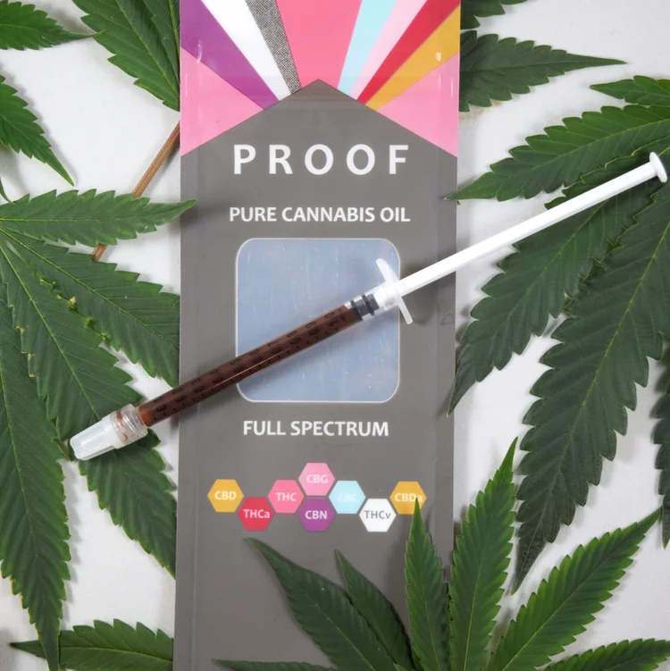 A syringe filled with dark cannabis oil rests on packaging labeled "Proof Pure Cannabis Oil Full Spectrum," surrounded by green cannabis leaves. The packaging highlights cannabinoids like CBD, THC, CBG, CBN, and THCV. CA Norml