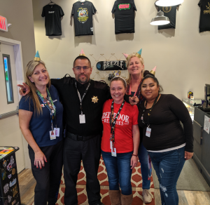 Five adults, four women and one man in a sheriff uniform, stand smiling indoors at Red Door Remedies. All wear paper party hats and work badges. T-shirts hang on the wall behind them, and a patterned carpet is on the floor. CA Norml