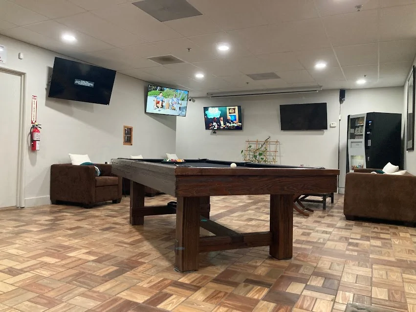 A recreation room at Double Eye Dispensary features a pool table in the center, three wall-mounted TVs, brown armchairs with white pillows, a vending machine, and a fire extinguisher. The floor is covered with patterned tiles for a cozy dispensary lounge vibe. CA Norml