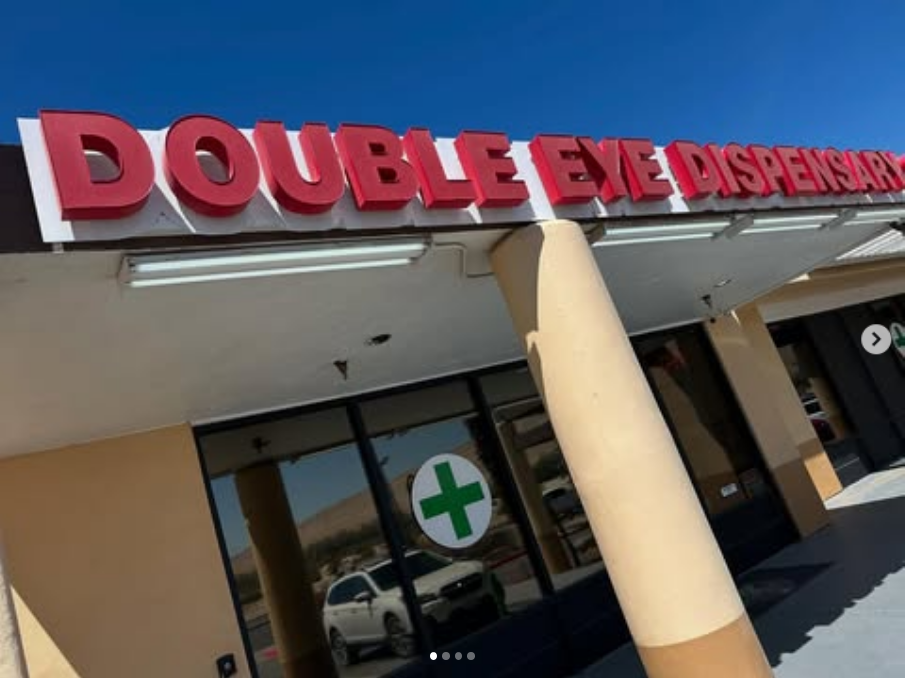 A storefront with large red letters reading “DOUBLE EYE DISPENSARY” above the entrance welcomes visitors. The door features a green medical cross symbol, while the sunny sky hints at a relaxing Dispensary Lounge experience inside. CA Norml