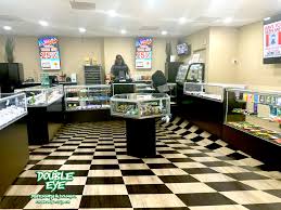 A brightly lit Double Eye Dispensary interior with glass display cases showcasing products, a black-and-white checkered floor, and a person behind the counter. Colorful signs adorn the back wall, creating a welcoming cannabis lounge vibe. CA Norml