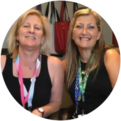 Two women smiling and sitting close together, both wearing black tops and colorful Red Door Remedies lanyards around their necks. Handbags are visible hanging in the background. CA Norml