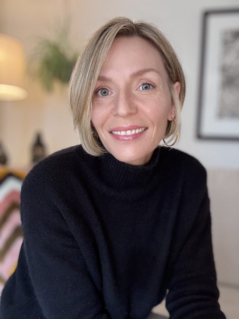 A woman with short blonde hair and blue eyes smiles at the camera. She is wearing a black turtleneck sweater and is indoors, with blurred decor visible in the background. CA Norml