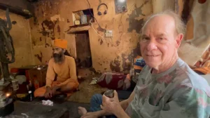 Ed Rosenthal holding a metal cup sits on the floor of a rustic, weathered room next to a man in orange robes and a turban. The room has peeling paint, shelves, and various items scattered around. Ca NORML