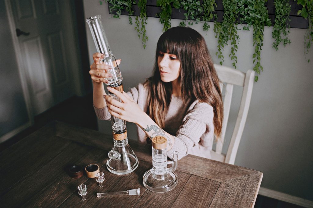 A woman with long brown hair assembles a glass bong at a wooden table, with various bong components laid out in front of her. Green plants hang on the gray wall behind her. Ca NORML