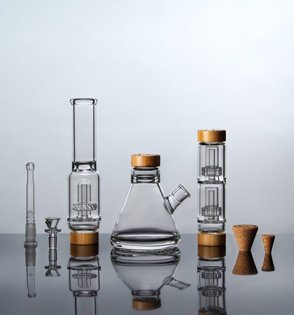 A set of glass smoking accessories with cork lids and bases, including a beaker-shaped water pipe, two tall cylindrical attachments, a glass bowl, and two cork stoppers, all arranged on a reflective surface. Ca NORML