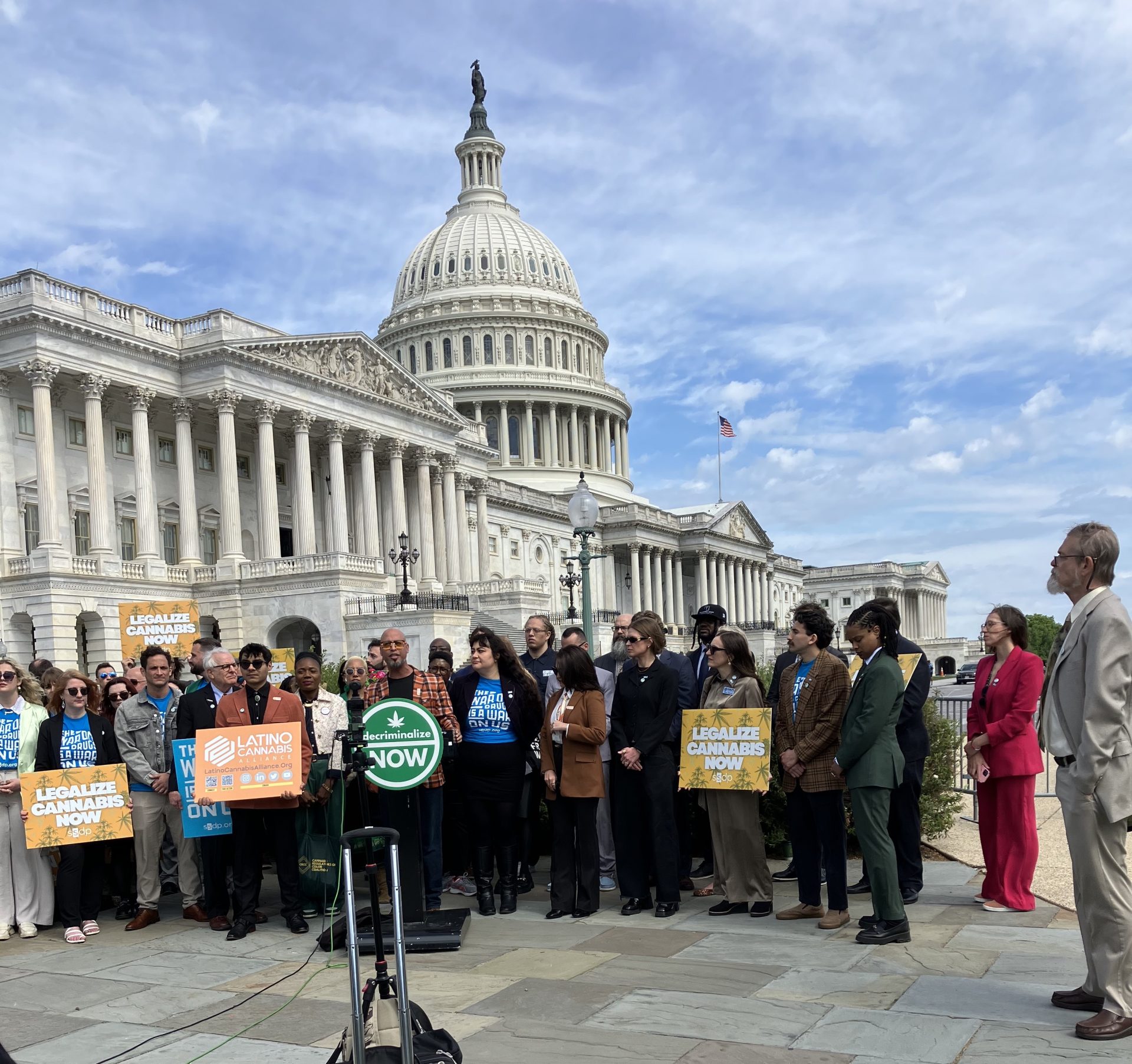 A diverse group gathers in front of the U.S. Capitol building for Lobby Day, holding signs supporting cannabis legalization and Latino representation, during an outdoor press event organized by the Cannabis Unity Coalition on a sunny day. Ca NORML