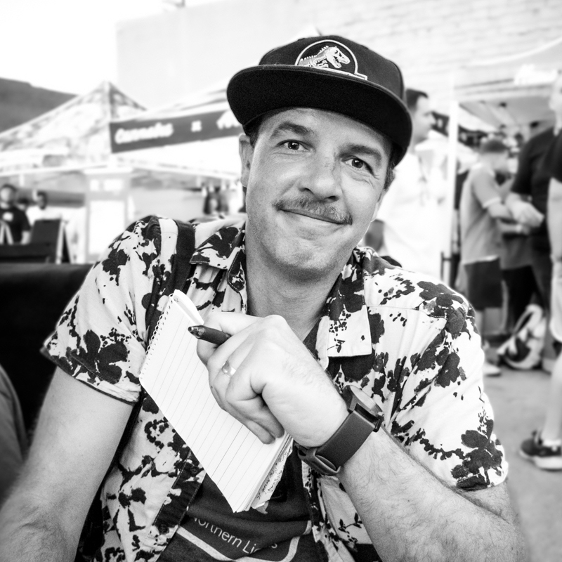 David Downs, in a patterned shirt and cap, sits outdoors holding a notepad and pen, smiling at the camera. People and tents are visible in the blurred background. The black-and-white photo presents a candid moment. Ca NORML