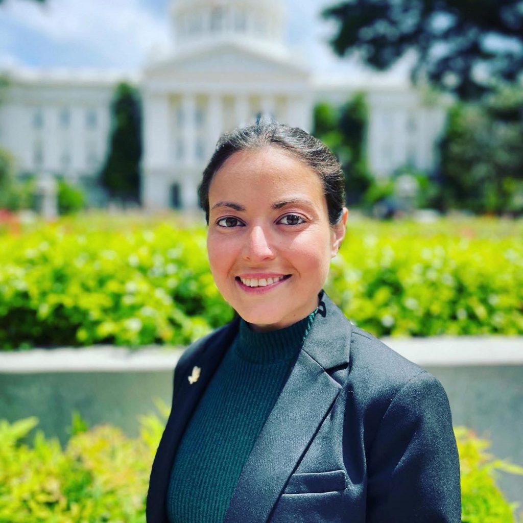 Lauren Mendelsohn, wearing a dark blazer and green turtleneck, stands smiling outdoors in front of a blurred white building with columns and a dome, surrounded by greenery and bright sunlight. Ca NORML